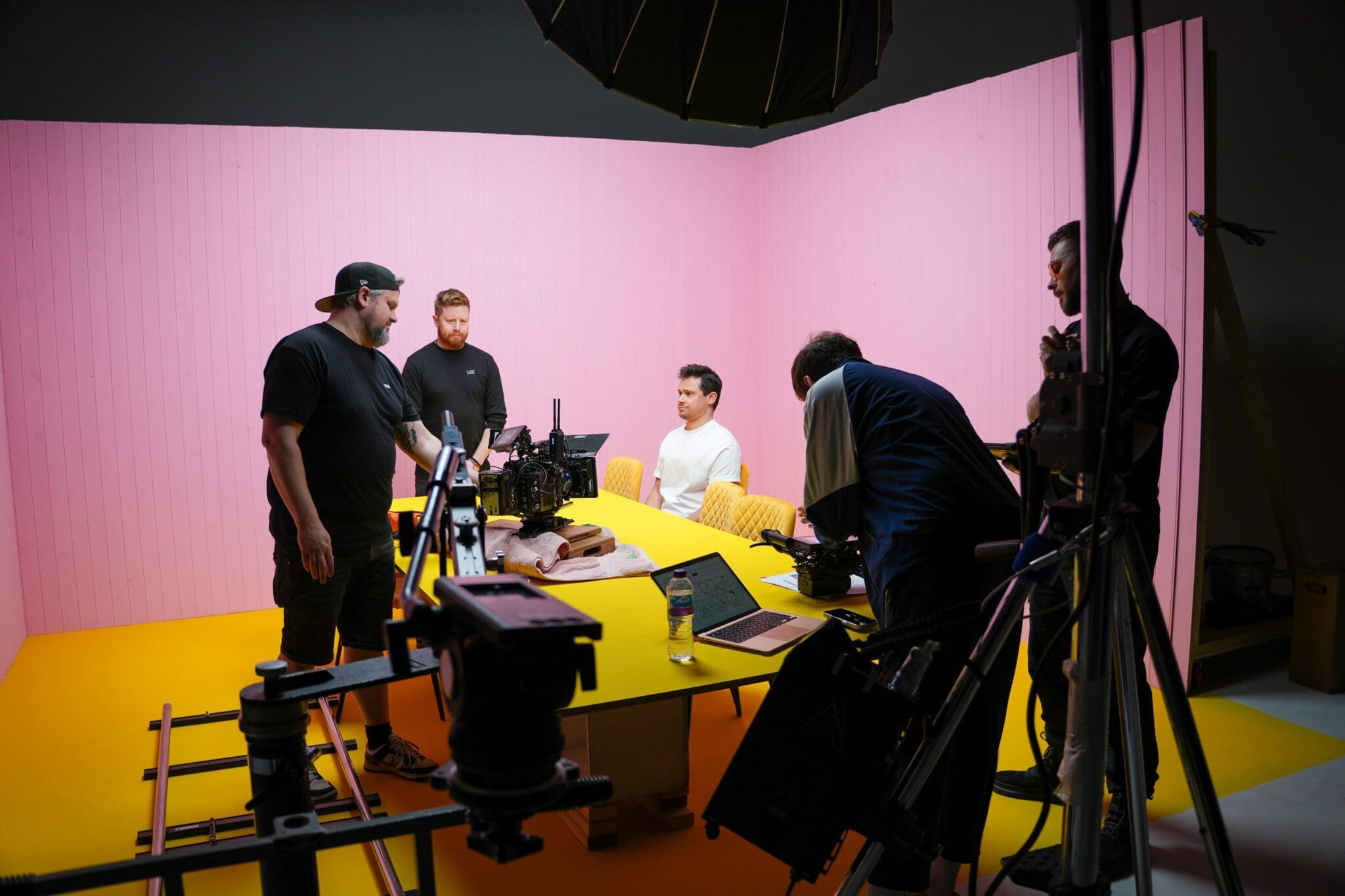 Five people work on a film or video set with cameras and equipment around a yellow table, against a pink wall and yellow floor backdrop.