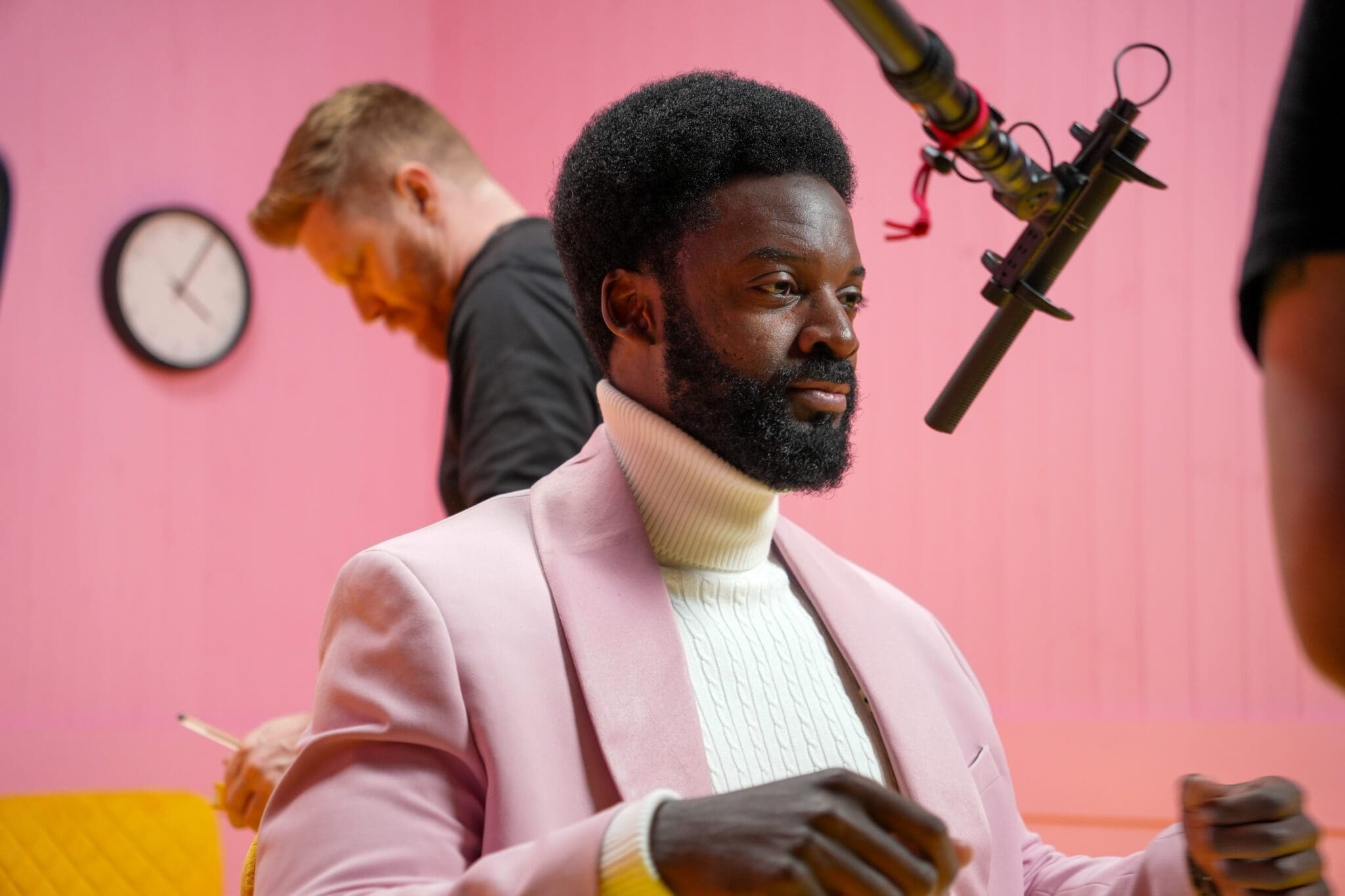 A man in a pink suit and white polo neck sits under a boom microphone in a studio, while another man writes in the background; the wall is pink with a clock.
