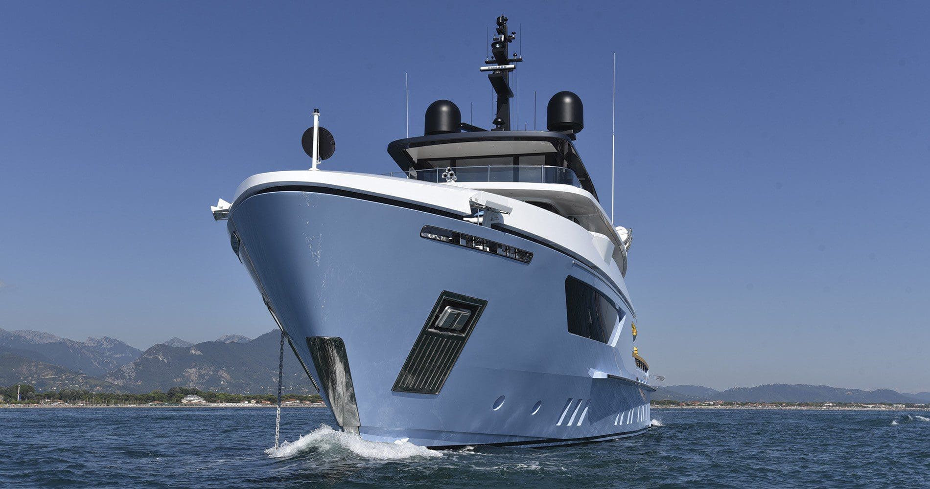 A large modern yacht with a blue hull is cruising on calm water with mountains and a clear sky in the background.