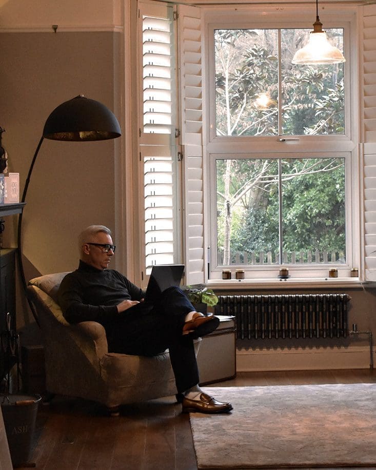 A person sits in an armchair using a laptop near a large window with shutters, in a lounge with wooden floors and modern decor.