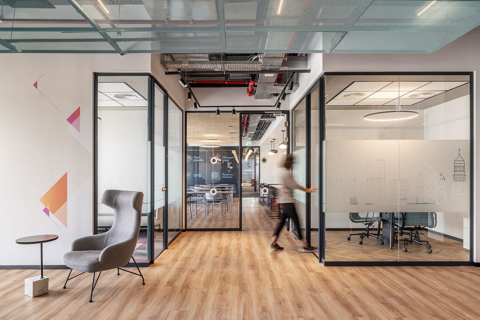 Modern office interior with glass-walled meeting rooms, wooden flooring, a grey armchair, and a person walking through a glass door.