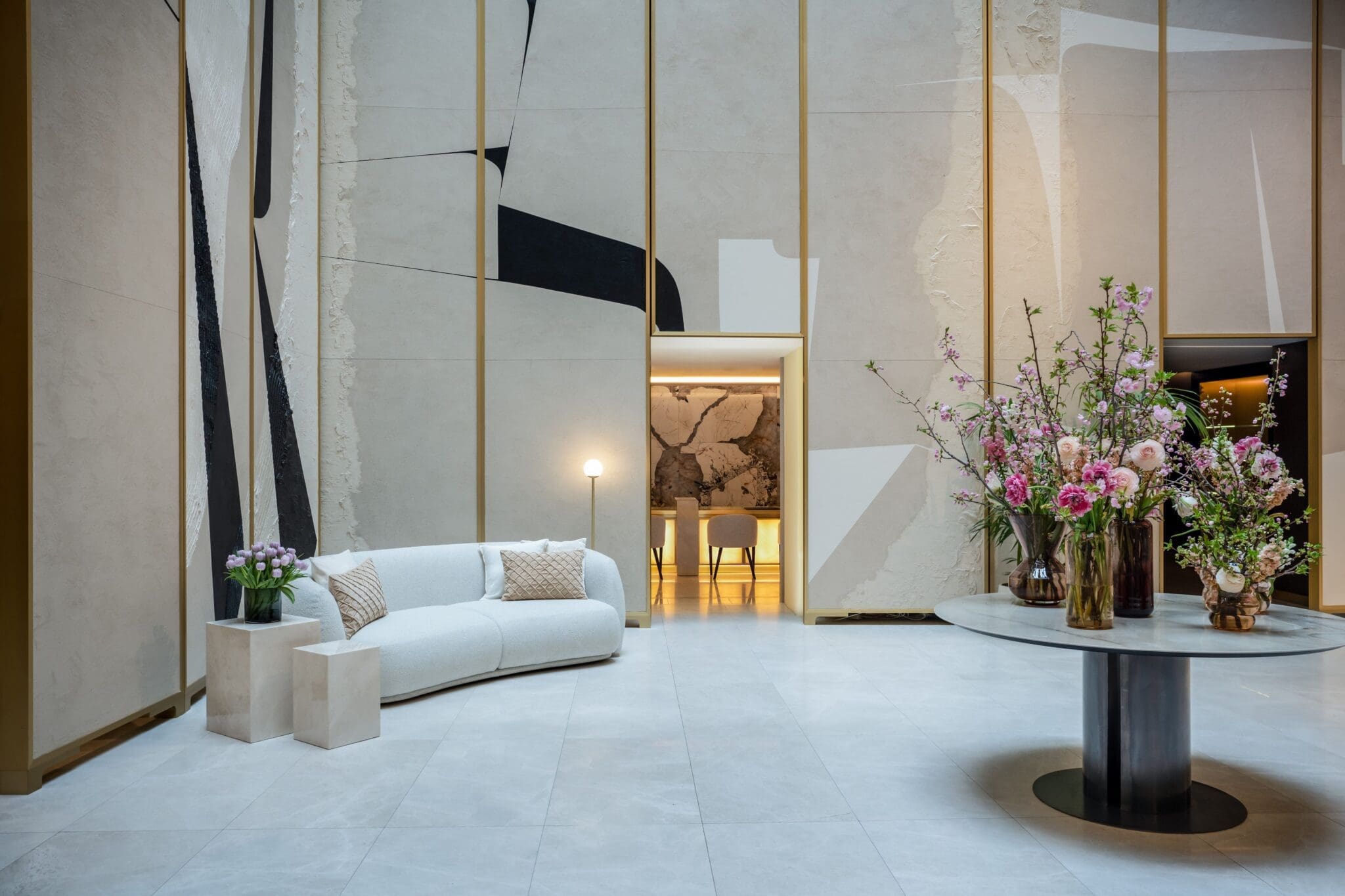 Modern lobby with a white curved sofa, flower arrangements on a round table, and abstract wall art; a lit room is visible in the background.
